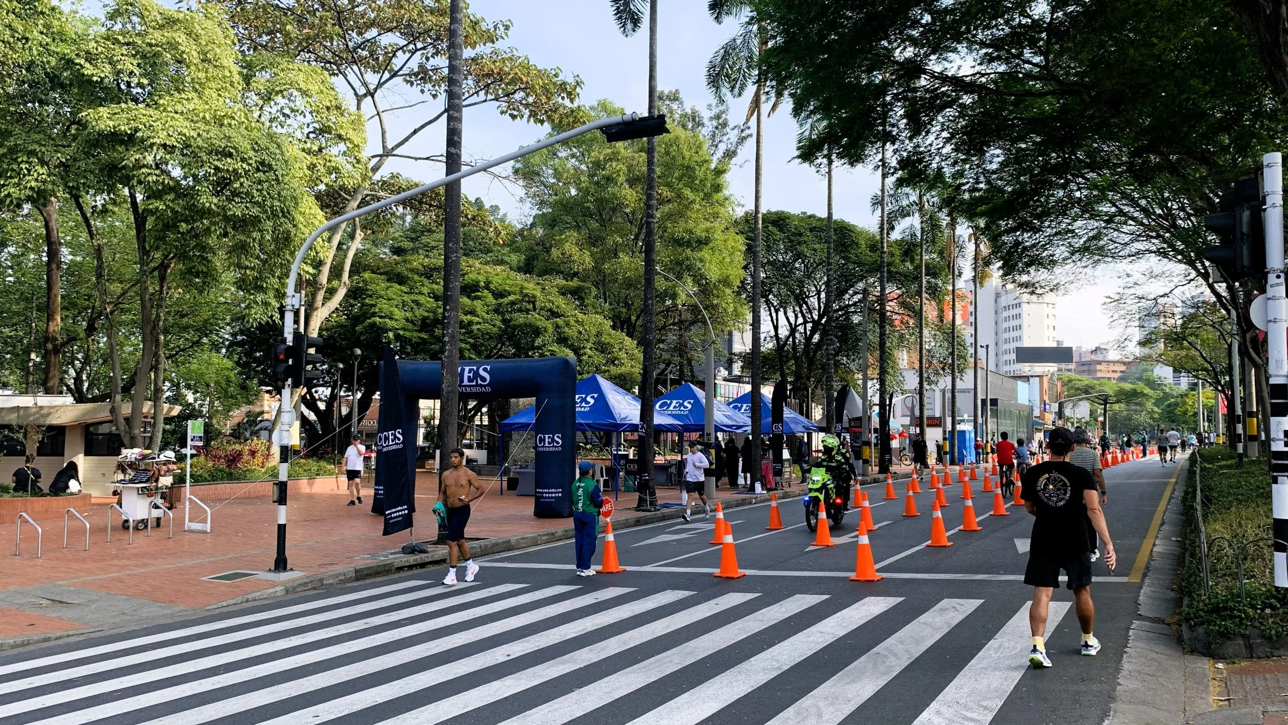 Participantes de una jornada de actividad física utilizan una vía urbana temporalmente adecuada para el evento. En el recorrido se observan personas caminando, corriendo y desplazándose en bicicleta, junto a puntos de apoyo identificados con la imagen institucional de la Universidad CES.