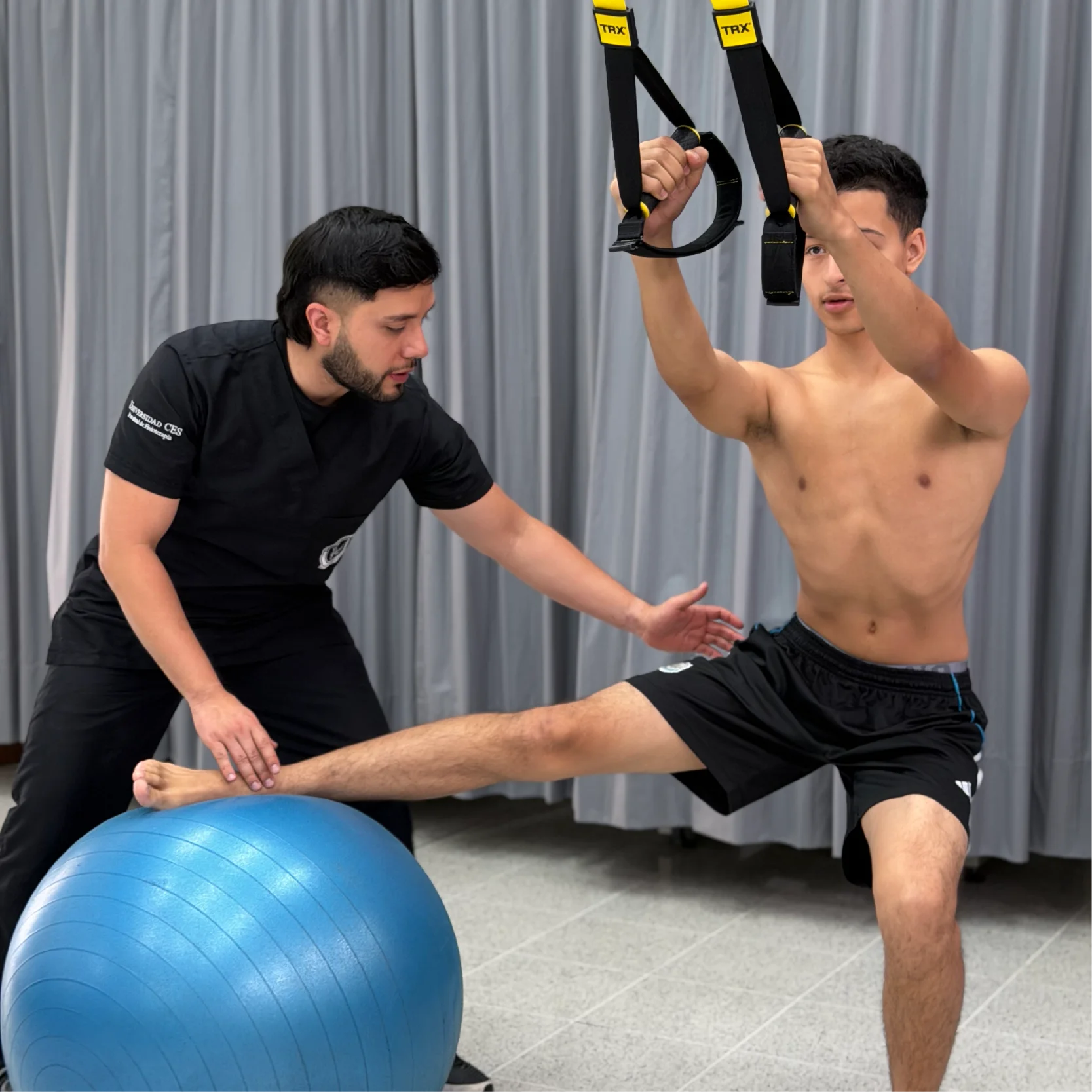 Persona realiza un ejercicio de equilibrio y estabilidad utilizando bandas de suspensión y una pelota de ejercicio, mientras otra persona supervisa y guía la actividad en un espacio de entrenamiento y rehabilitación física.