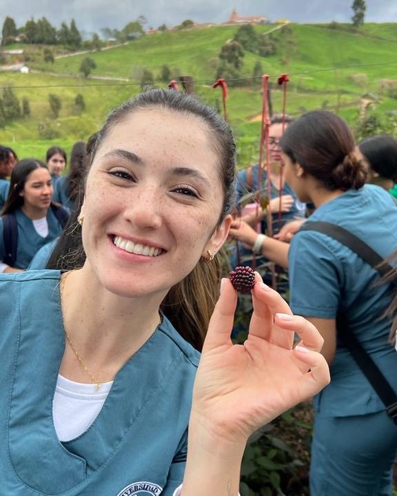 Persona con uniforme clínico sostiene una mora en la mano durante una actividad al aire libre, con otras personas y vegetación en el fondo.