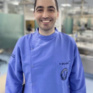 Persona con uniforme clínico azul de pie en un entorno odontológico, con equipos y monitores al fondo.