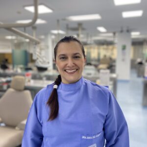 Persona con uniforme clínico azul de pie en un entorno odontológico, con sillones y equipos visibles al fondo.