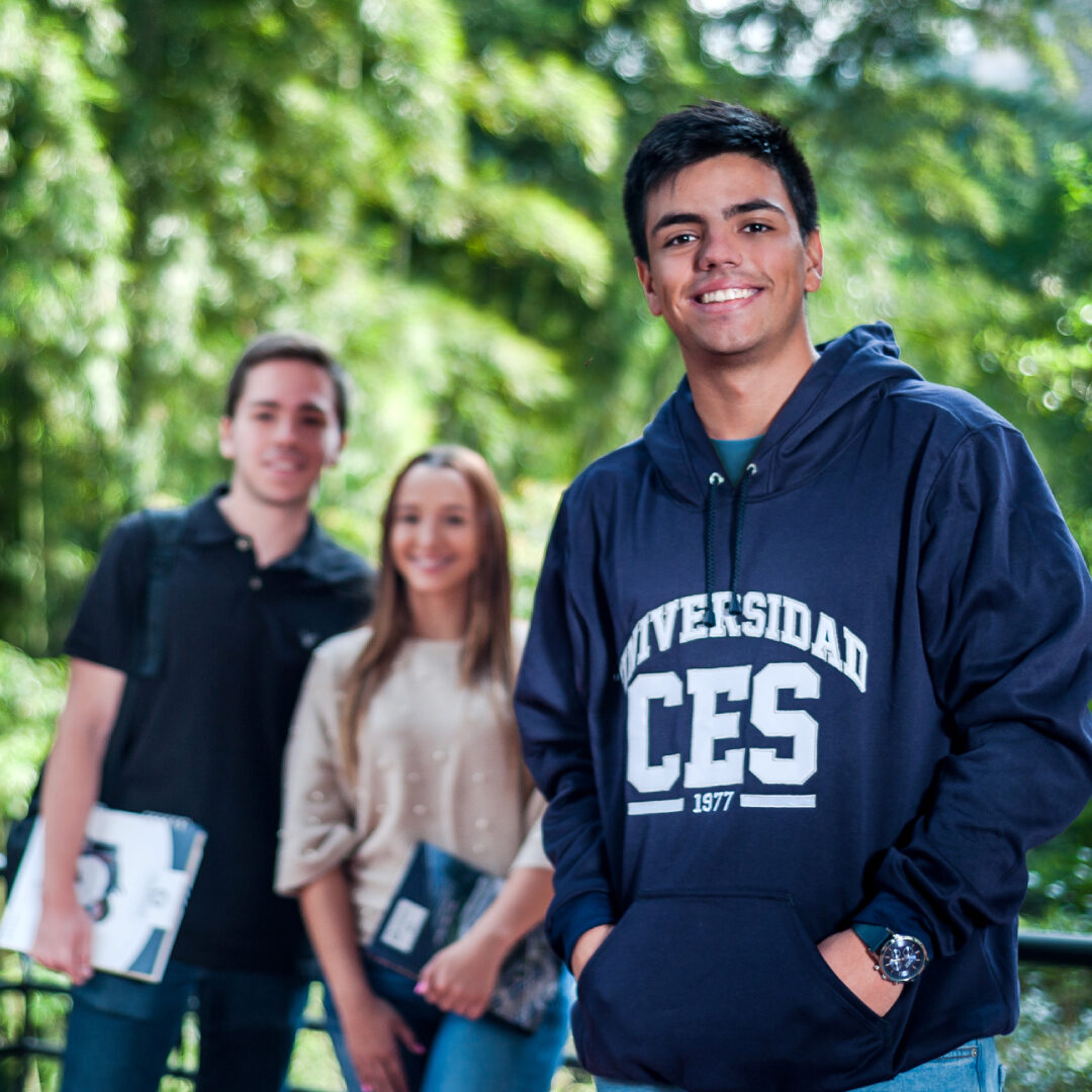 Grupo de tres personas de pie al aire libre; una en primer plano con sudadera azul marino que dice ‘Universidad CES’, y dos detrás sosteniendo cuadernos