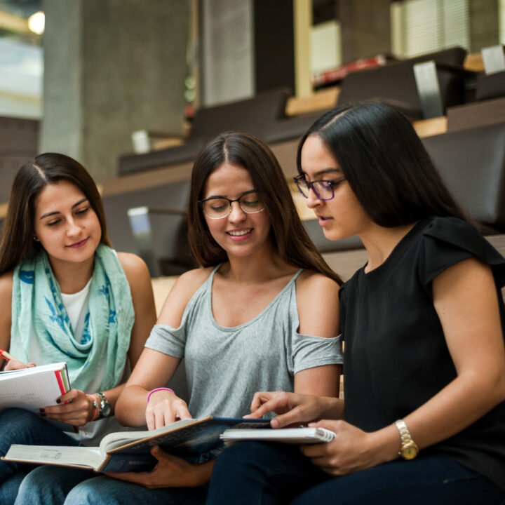 Tres personas sentadas en gradas interiores revisan libros y cuadernos abiertos.
