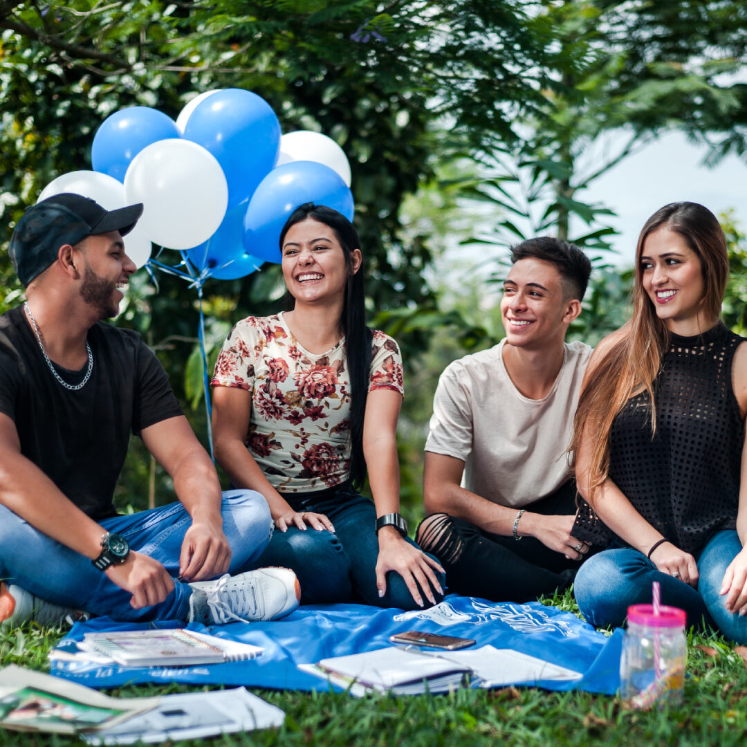 Grupo de personas sentadas sobre una manta azul en un área verde, con globos blancos y azules detrás y materiales impresos distribuidos en el suelo.