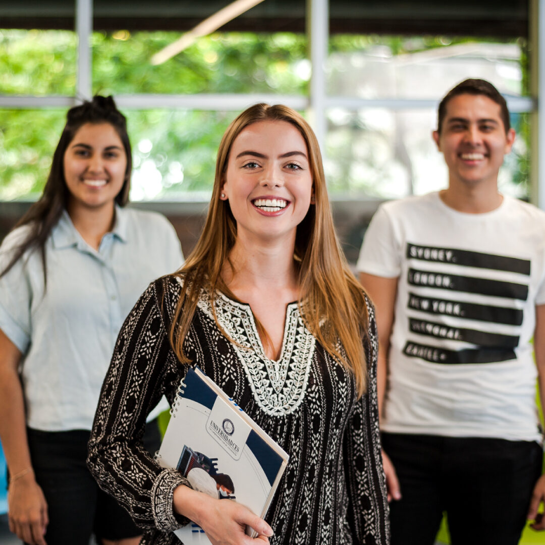 Tres personas están de pie en un espacio interior iluminado, con una ventana grande al fondo. La persona en primer plano sostiene una carpeta, mientras las otras dos están ligeramente detrás.