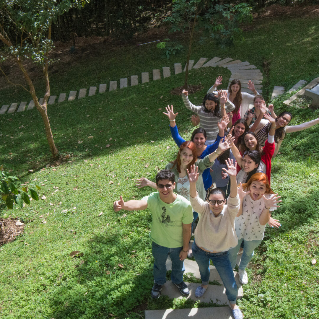 Grupo de estudiantes en un jardín al aire libre, alineadas sobre un sendero de piedra, levantando los brazos en señal de saludo. Al fondo se observan árboles y césped.