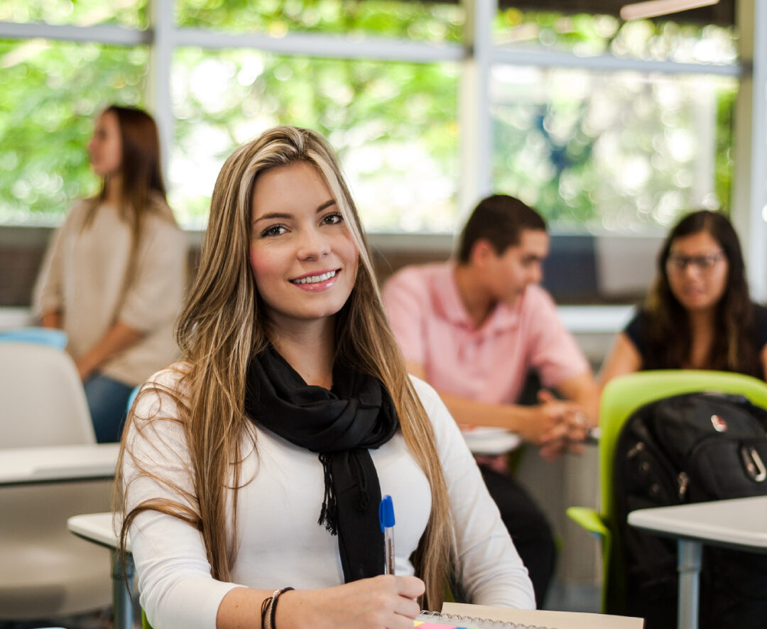 Estudiante sentada en un aula con cuadernos y bolígrafo sobre la mesa, mientras otras personas están en segundo plano. El espacio tiene ventanales amplios que dejan entrar luz natural y muestran vegetación exterior.