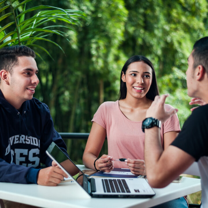 Grupo de tres personas sentadas alrededor de una mesa al aire libre, conversando y trabajando con una computadora portátil y una tableta