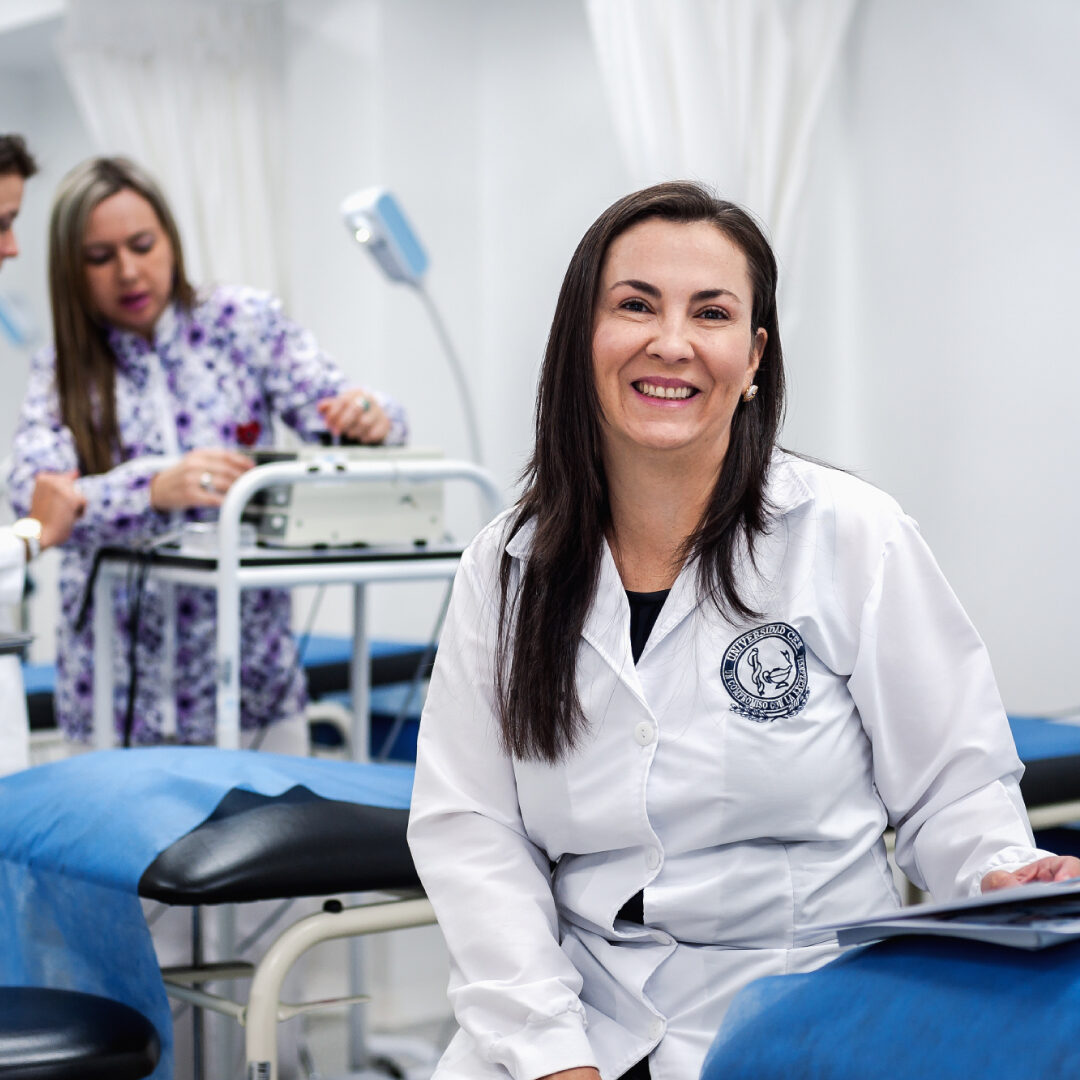 Persona con bata blanca y emblema institucional está sentada en una camilla en un consultorio, mientras otra persona manipula un equipo médico en el fondo.