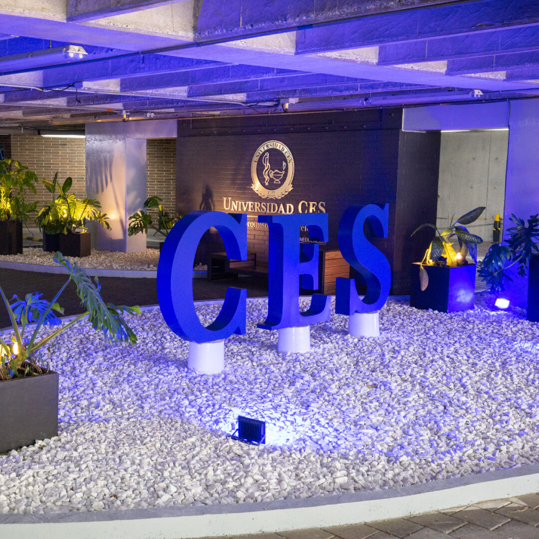Entrada principal de la Universidad CES con un letrero grande en color azul que dice “CES” sobre un área cubierta de piedras blancas decorativas. Al fondo se observa una pared con el logotipo y el texto “Universidad CES”, rodeada de plantas y luces ambientales en tonos azules y cálidos.