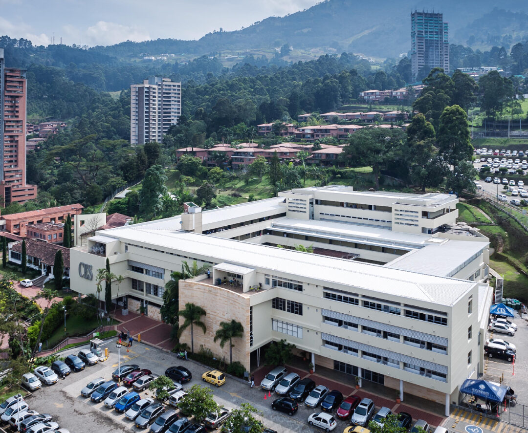 Vista aérea de un edificio blanco de varios pisos rodeado de zonas verdes y estacionamiento con numerosos vehículos. Al fondo se observan edificios residenciales, árboles y montañas.