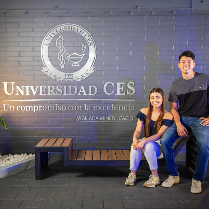 Dos personas sentadas en un banco frente a una pared con el logo y el nombre ‘Universidad CES’ y el texto ‘Un compromiso con la excelencia’.