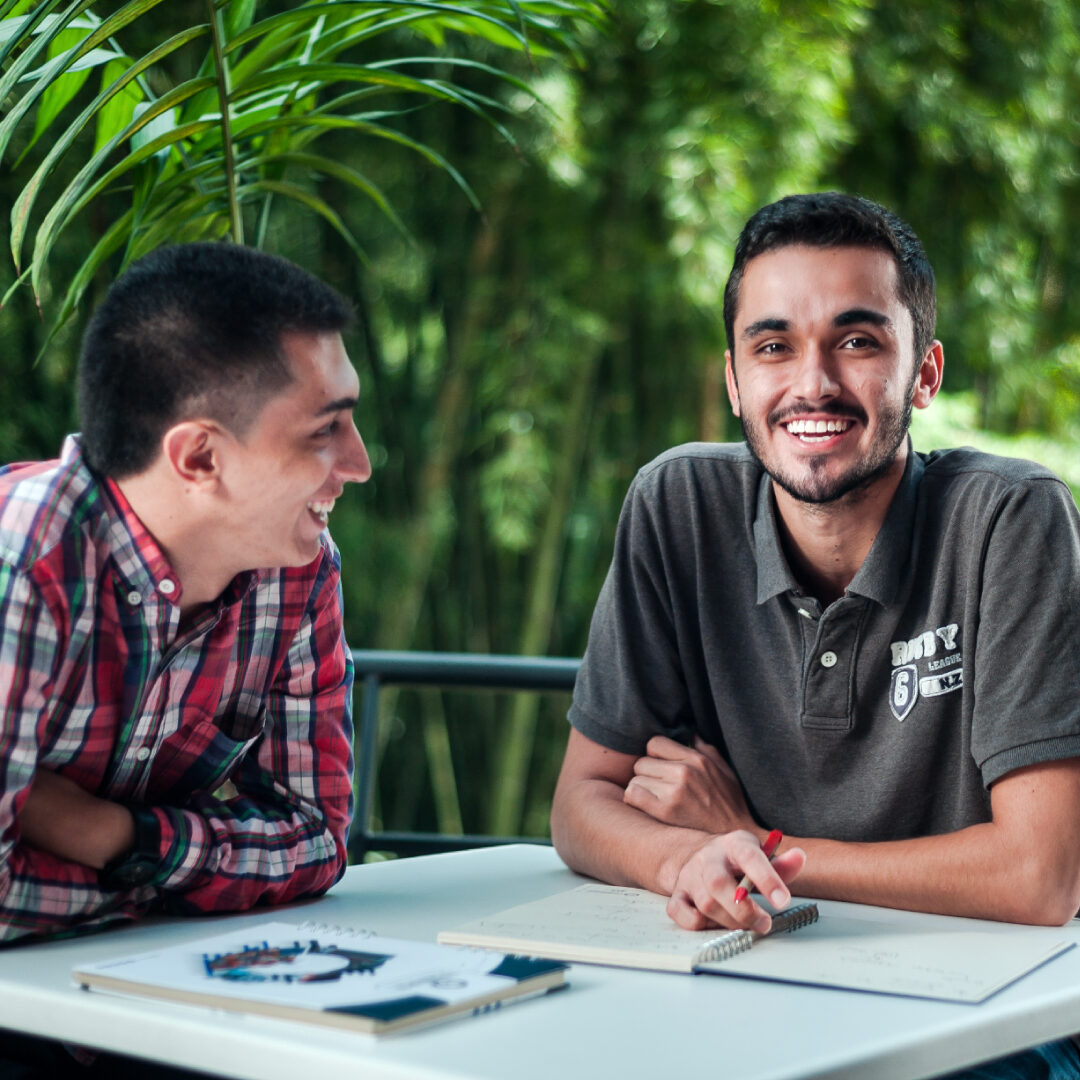 Dos personas sentadas en una mesa al aire libre, conversando y revisando documentos, con vegetación en el fondo.