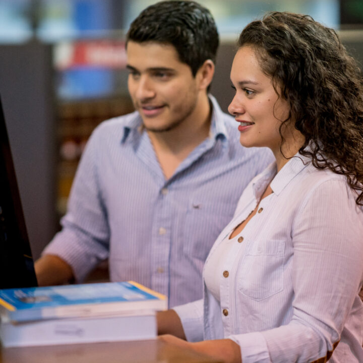Dos personas están de pie frente a un monitor, con libros y documentos sobre una mesa en una biblioteca.