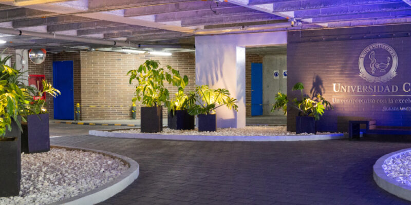 Zona interior amplia con piso oscuro y techo de concreto, decorada con plantas en macetas negras sobre áreas de piedras blancas; al fondo hay una pared azul con el logo "Universidad CES" iluminada con luces azules y violetas.
