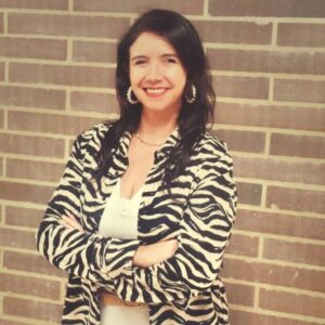 Persona de pie frente a una pared de ladrillos, con los brazos cruzados, vistiendo una blusa blanca y una chaqueta con estampado de rayas tipo cebra.