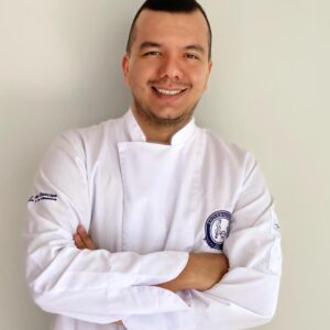 Persona de pie frente a una pared clara, usando chaqueta blanca tipo uniforme profesional con logotipo bordado en el pecho y brazos cruzados.