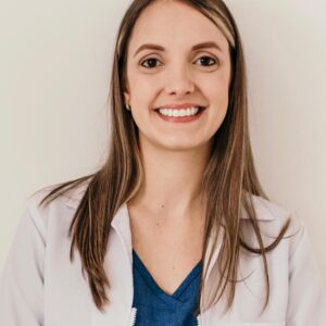 Persona de pie frente a una pared clara, usando chaqueta blanca abierta sobre una prenda azul tipo uniforme.