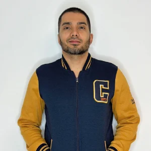 Persona de pie frente a una pared blanca, usando chaqueta en color azul marino con mangas mostaza y detalles en puños y cuello; en el pecho se observa una letra ‘C’ bordada.