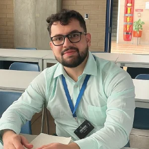 Persona sentada en un aula, con camisa clara y gafete colgado al cuello, sosteniendo documentos sobre una mesa.