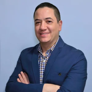 Persona de pie frente a una pared azul, usando saco formal color azul marino sobre camisa de cuadros en tonos azul y blanco, con los brazos cruzados.