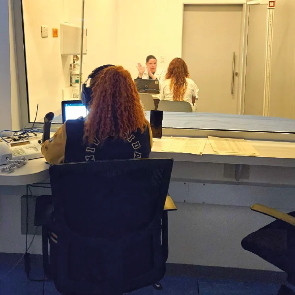 Persona con audífonos y computadora observa a través de un vidrio a dos individuos en una sala, participando en lo que parece ser una sesión de evaluación o práctica supervisada.