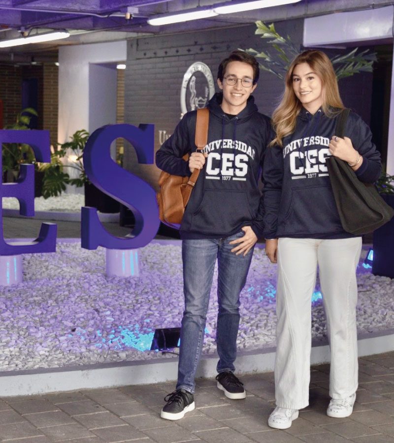 Dos personas están de pie junto a letras grandes que forman parte de la palabra “CES”, vistiendo sudaderas con el texto “Universidad CES” y llevando bolsos. El entorno es un espacio interior con decoración moderna y plantas.