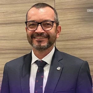 Persona vestida con traje formal oscuro, camisa blanca y corbata azul con pequeños detalles rojos, de pie frente a una pared con acabado de madera.