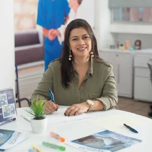 Persona sentada en una oficina, escribiendo sobre documentos en una mesa blanca que tiene una planta pequeña, resaltadores y revistas; viste camisa verde oliva y lleva reloj en la muñeca.