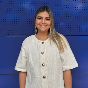 Persona de pie frente a un fondo azul, usando una chaqueta o blusa clara de manga corta con botones grandes dorados en el centro.