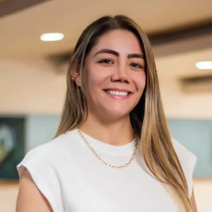 Persona de pie en un espacio interior iluminado, usando blusa sin mangas de color blanco y un collar dorado sencillo; el fondo muestra paredes claras y luces circulares en el techo.