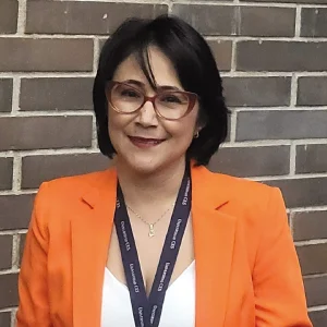 Persona de pie frente a una pared de ladrillos, usando chaqueta naranja sobre una prenda blanca y un cordón oscuro con gafete colgado al cuello.