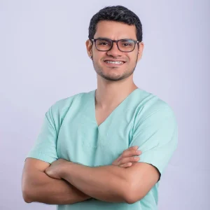 Persona con cabello corto y oscuro, vistiendo camiseta tipo clínica color verde claro, con los brazos cruzados, frente a un fondo liso de tono claro.