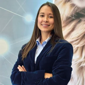 Persona con cabello lacio y largo, vistiendo camisa formal azul claro y saco azul oscuro, con los brazos cruzados, frente a un fondo con diseño gráfico y textura difusa.