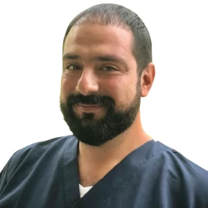 Persona con cabello corto y barba, vestida con uniforme azul oscuro tipo clínico sobre camiseta blanca, en fondo blanco.