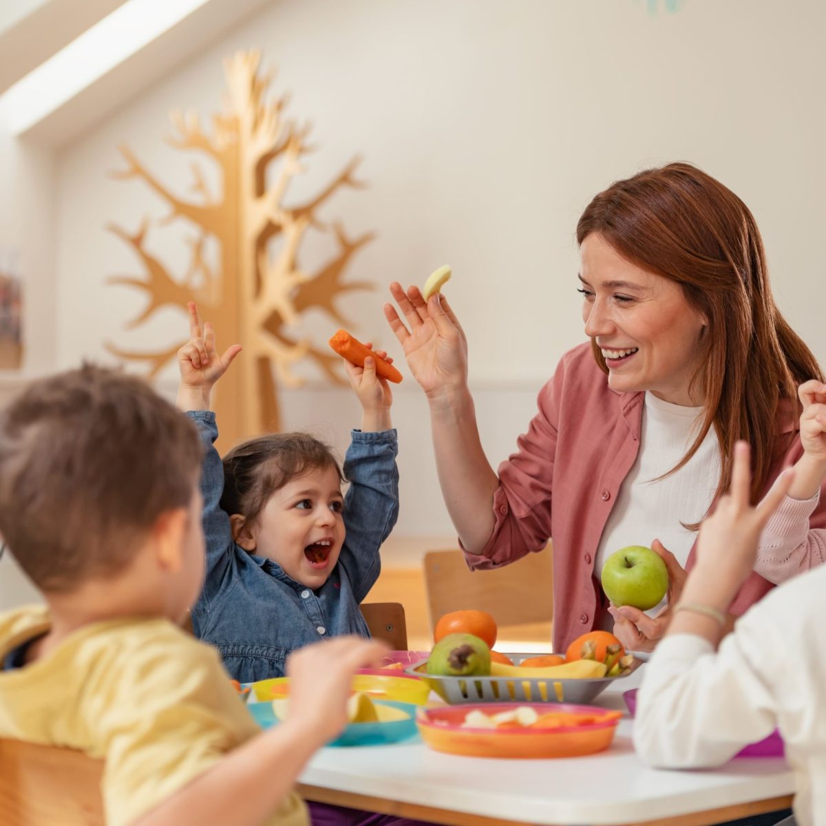 Grupo de niños sentados alrededor de una mesa con frutas, levantando las manos junto a una persona adulta en un ambiente educativo.