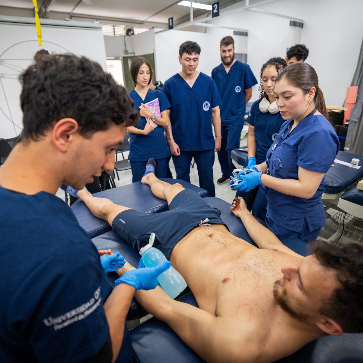 Grupo de personas con uniformes clínicos realiza una práctica médica en un entorno educativo; una persona está acostada en una camilla mientras otras aplican gel y utilizan un dispositivo de ultrasonido en la zona abdominal.