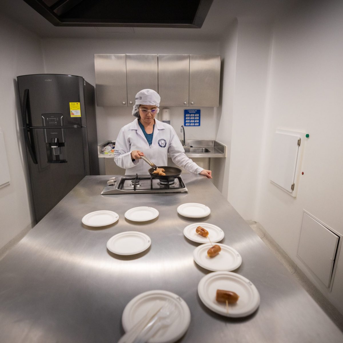 Persona con uniforme y gorro de cocina prepara alimentos en una estufa dentro de una cocina industrial; sobre la mesa metálica hay varios platos blancos con porciones servidas.