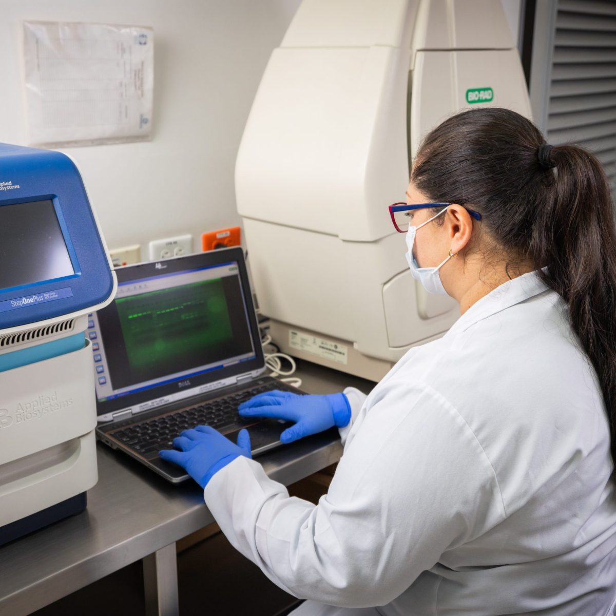 Persona con bata blanca y guantes azules trabaja en un laboratorio, utilizando un computador portátil conectado a equipos especializados para análisis biomédico o genético.