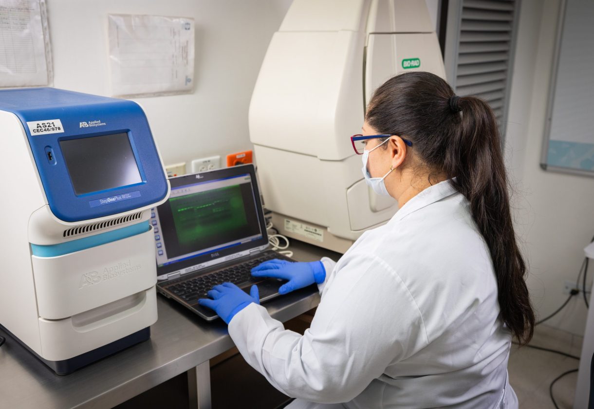 Persona con bata blanca y guantes azules trabaja en un laboratorio, utilizando un computador portátil conectado a equipos especializados para análisis biomédico o genético.