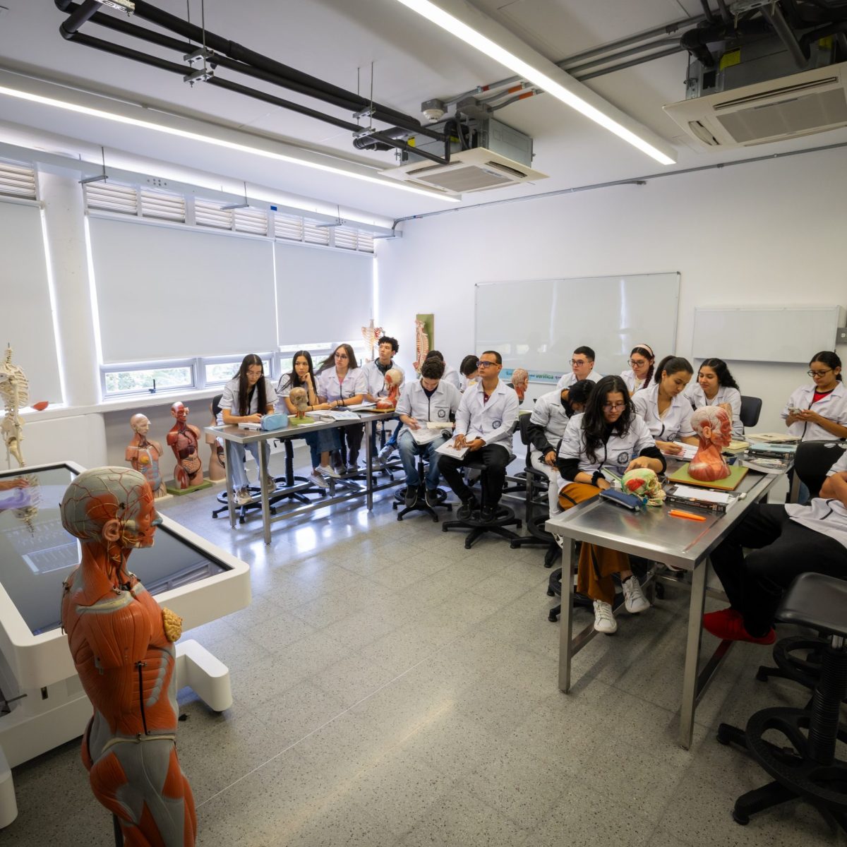 En un aula equipada para enseñanza médica, una persona explica el uso de una mesa anatómica digital mientras un grupo de estudiantes con batas blancas observa; sobre la mesa cercana hay modelos anatómicos y un maniquí humano.