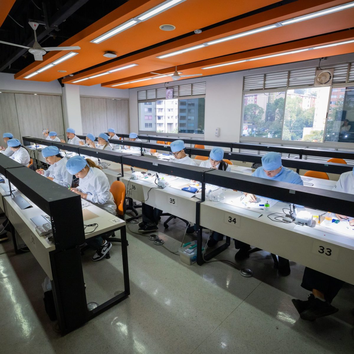 Varias personas con uniformes clínicos y gorros trabajan en estaciones individuales dentro de un aula equipada, realizando prácticas sobre materiales odontológicos en un entorno educativo.