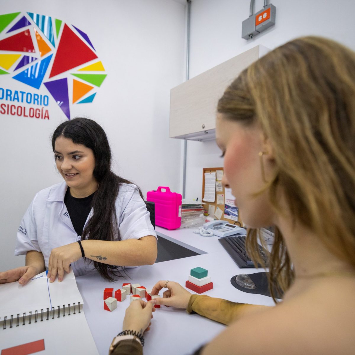 Dos personas están sentadas en un escritorio dentro de un laboratorio de psicología; una sostiene un cuaderno abierto mientras la otra manipula bloques de diferentes formas y colores sobre la mesa.