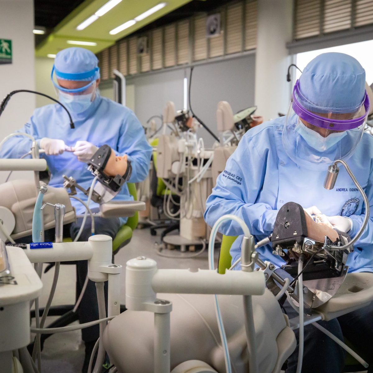 Dos personas con indumentaria odontológica y guantes realizan prácticas sobre simuladores dentales en un entorno clínico equipado con instrumental y materiales odontológicos.