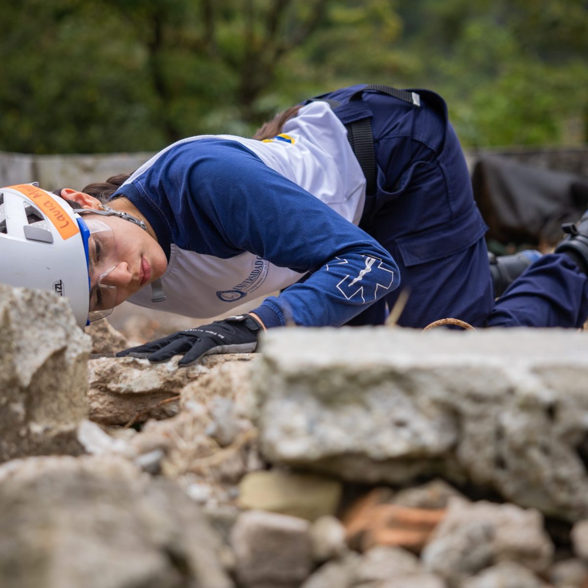 Persona con casco y guantes revisa cuidadosamente escombros en una zona al aire libre, posiblemente en labores de búsqueda o rescate.