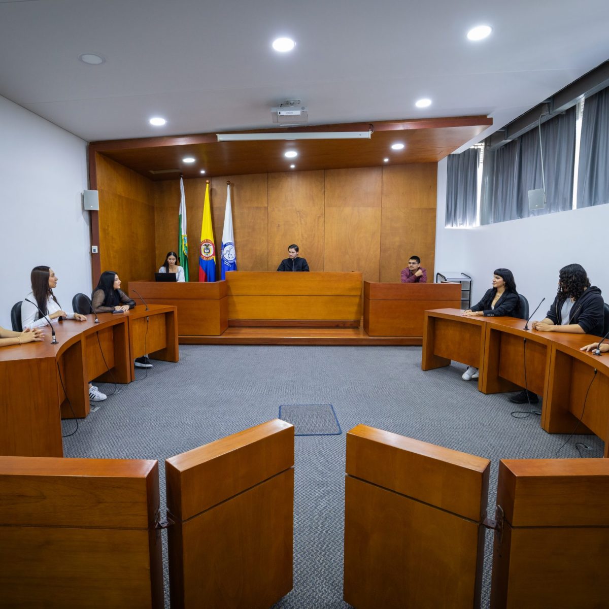 Varias personas están sentadas en escritorios dispuestos en forma de U dentro de una sala que simula un tribunal; al frente, una persona está ubicada en el estrado central con banderas detrás, indicando un escenario de práctica jurídica o audiencia simulada.