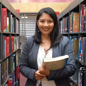 Persona de pie en un pasillo de biblioteca, sosteniendo un libro abierto con ambas manos; viste chaqueta gris sobre blusa blanca y está rodeada de estanterías llenas de libros.