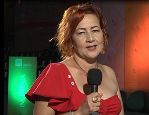 Persona con vestido rojo sostiene un micrófono en un espacio interior con fondo oscuro y paneles iluminados