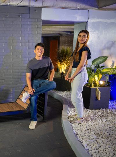 Dos personas en un espacio interior con decoración moderna, una sentada en un banco y otra de pie junto a un área con piedras blancas y plantas iluminadas.