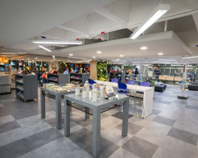 Espacio interior amplio con mesas y estanterías, iluminación artificial y decoración moderna, similar a una zona de estudio o biblioteca.